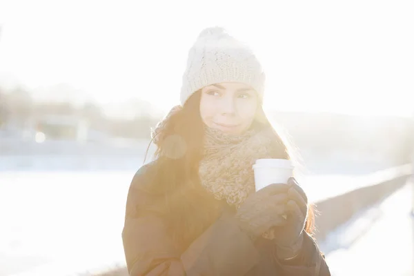 Cute brunette chick enjoying hot drink.White paper takeaway cup from local barista.Place your cafe logo on clean mug for refreshing drinks.Lens flare and bright sunlight.Model in warm winter wear