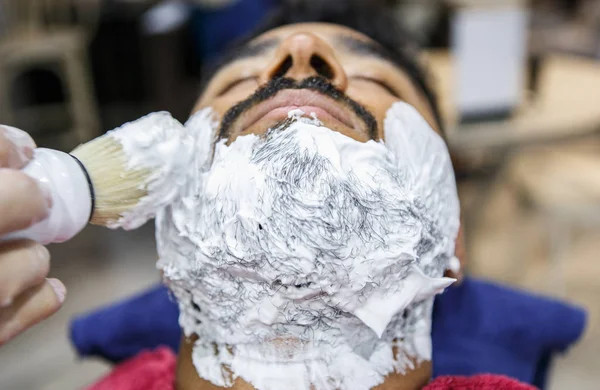 Barber spreads white shaving foam on client face with professional swob brush in barbershop salon.Hairdresser spreading cream on young black man beard before shave.Male beauty treatment concept