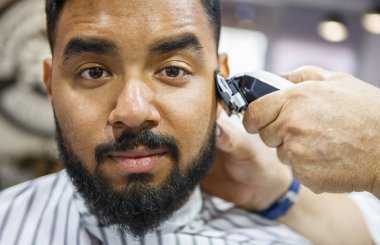 Portrait of young black man being clipped with professional clipper machine in barbershop.African guy makes new haircut and trim beard at barber salon. Male beauty treatment concept clipart