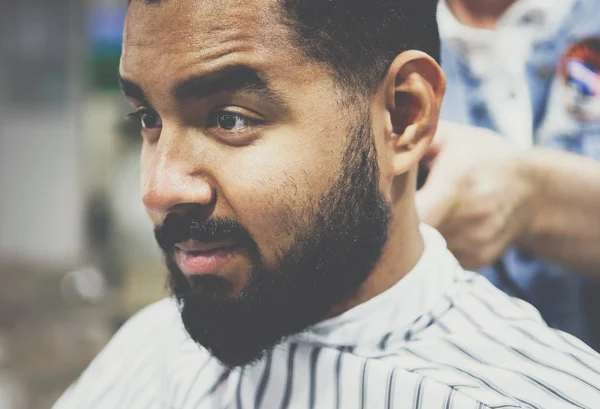 Portrait of unshaven young African man sitting in chair covered with blanket at barbershop salon while barber prepares his hair for cutting.Male beauty treatment process.Cut new haircut & trim beard