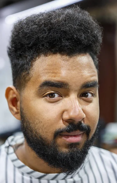 Portrait of young unshaven black man covered with blanket in barbershop salon waiting for new haircut and beard & mustache trimming. Male beauty treatment concept.