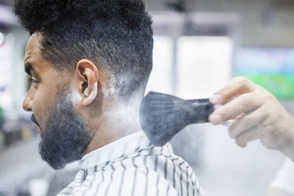 Barbers hand spread talcum powder on clients neck with professional shaving brush in barbershop salon.Male beauty treatment concept.Young black man getting new haircut in barber shop studio