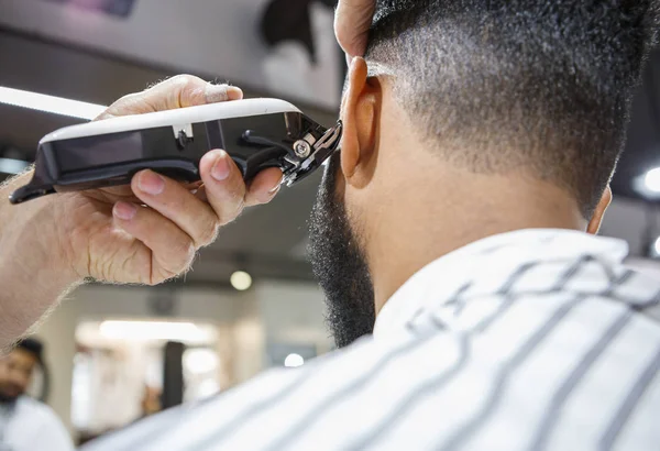 Barber trim hair with clipper on young unshaven black man in barbershop studio.Professional hairdresser cut hair with electric shearer machine on African guy. Male beauty treatment process