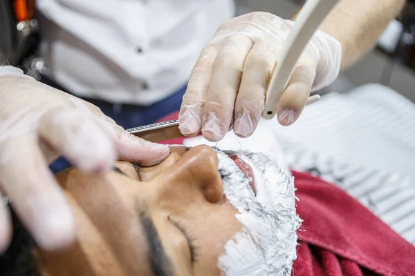 Barber cut clients beard with sharp razor shaver in barbershop.Young black man with white shaving foam on face get new haircut in salon. Male beauty treatment concept