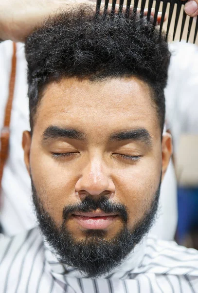 Portrait of young black man getting new haircut in barbershop studio.Male beauty treatment concept.Handsome African guy sitting in barber salon while hairdresser brushes his hair with comb tool