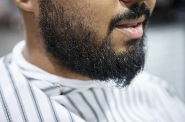 Genç adam onun saç kesme Kuaför bekler tıraşsız sakal ile siyah. İstemci berber salonu sandalyede oturan battaniye ile kaplı. Erkek güzellik tedavi kavramı