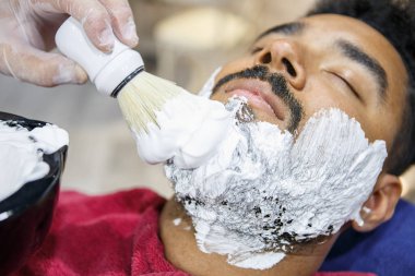 Berber berber salonu çubukla fırça ile genç siyah adam yüzünde beyaz tıraş kremi yayılıyor. Portre erkek güzellik tedavi sürecinde