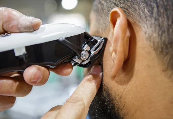 Barber trim African man client with clipper machine in barbershop.Professional trimmer tool cuts beard and hair on young black guy in barber shop salon. Male beauty treatment process in close up