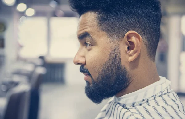 Portrait of handsome young black man with unshaven beard sitting in barbershop waiting for barber to make him pretty haircut and trim beard & mustache hair.Male beauty treatment concept