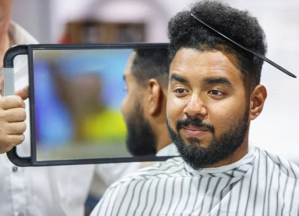 Portrait of black man with comb in curly hair looking in the mirror to the check haircut.Barber showing client his work in barbershop salon.Male beauty treatment concept