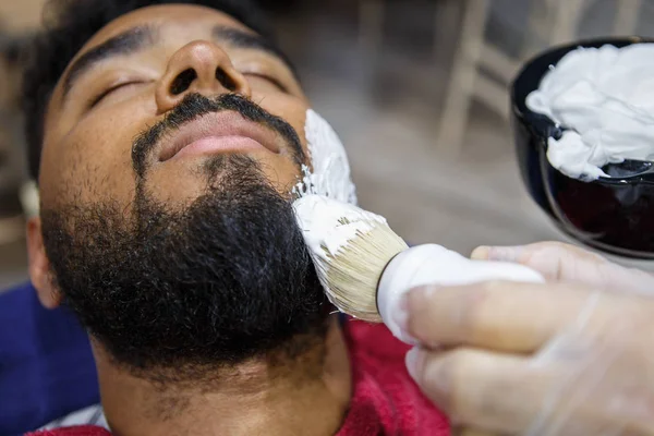 Barber spreads white shaving foam with professional brush on clients face for accurate shave.Young black man get his unshaven beard cut into shape in barbershop. Male beauty treatment concept