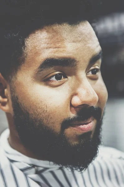 Handsome young black man with unshaven beard and mustache in barbershop waiting for barber hairdresser to cut his hair,trim mustache & beard.Male beauty treatment concept.Hairdresser client portrait