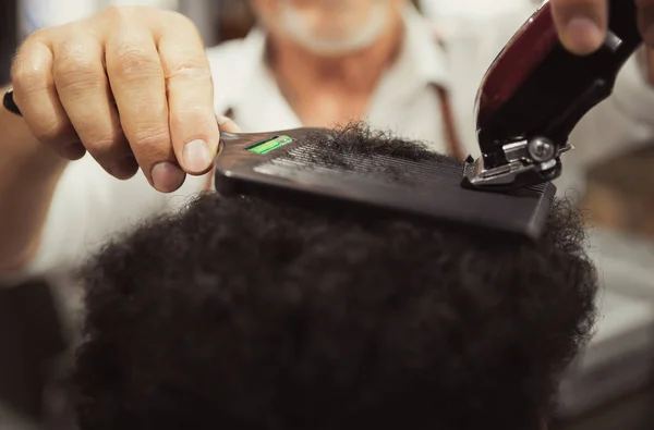 Barber shaves african male client with curly black hair using electric shaving machine in close up.Professional male stylist making new haircut to black guy in beauty salon.Beauty treatment for men