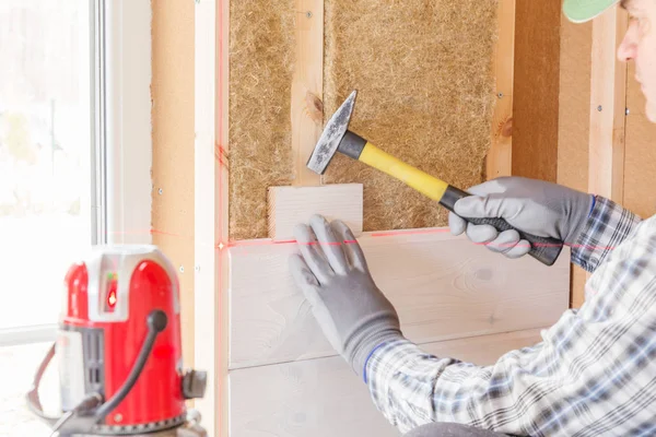The worker makes finishing works of walls with a white wooden board, using laser line level. Building heat-insulating eco-wooden frame house with wood fiber plates