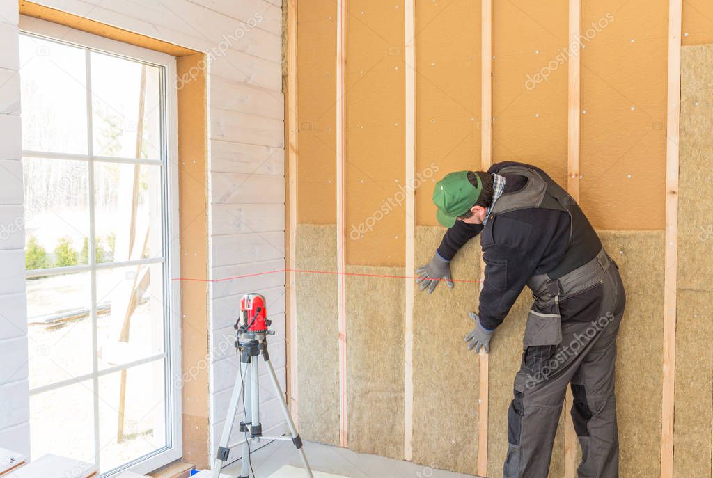 The worker makes finishing works of walls with a white wooden board, using laser line level. Building heat-insulating eco-wooden frame house with wood fiber plates