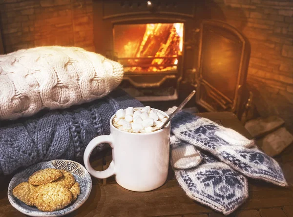 Stack of cozy knitted sweaters, handmade mittens and cup of coffee, cocoa, hot chocolate with marshmallows on old wooden table, near burning fireplace — Stock Photo, Image