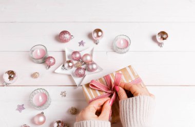 Noel paketleme işlemi sunar. Beyaz ahşap masa pembe süslemeleri, topları, mumlar, yıldız ile eller hediye pembe kurdele süsleyin. Yeni yıl, kış konsepti.