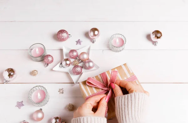 Proceso de embalaje regalos de Navidad. Mesa de madera blanca con decoraciones de color rosa, bolas, velas, estrellas, manos femeninas decorar regalo cinta rosa. Año nuevo, concepto de invierno . — Foto de Stock