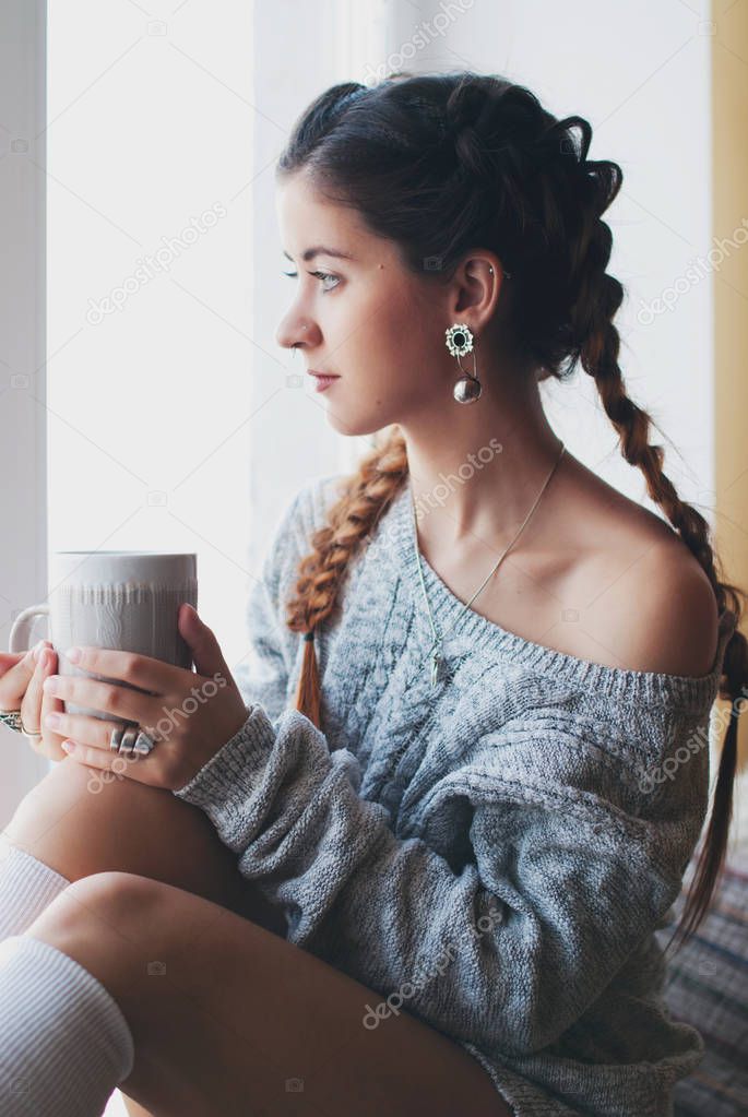 Young beautiful woman with mug of tea on the windowsill in the interior - sweet home concept