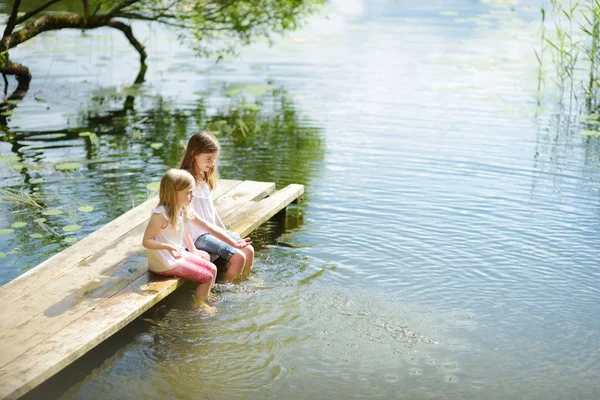 两个可爱的小女孩坐在河边的木台上 在温暖的夏日里把脚浸入水中 夏季家庭活动 — 图库照片