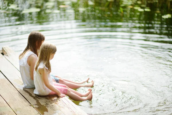 两个可爱的小女孩坐在河边的木台上 在温暖的夏日里把脚浸入水中 夏季家庭活动 — 图库照片