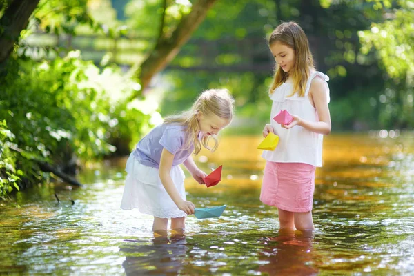 两个小妹妹在温暖晴朗的夏日里在河边玩纸船 孩子们在水里玩得很开心 幼儿暑期活动 — 图库照片