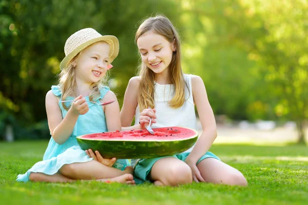 Two funny little sisters eating watermelon with the spoons outdoors on warm and sunny summer day. Healthy organic food for little kids.