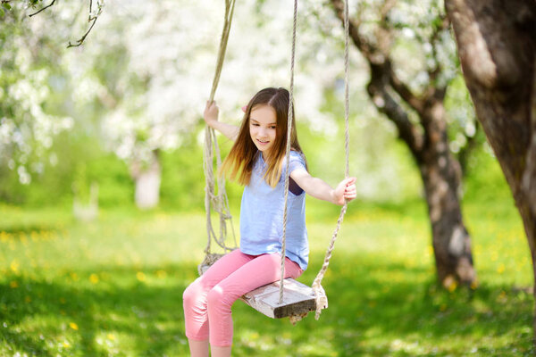 Милая маленькая девочка развлекается на качелях в цветущем старом яблоневом саду на открытом воздухе в солнечный весенний день. Весенние развлечения для детей
.