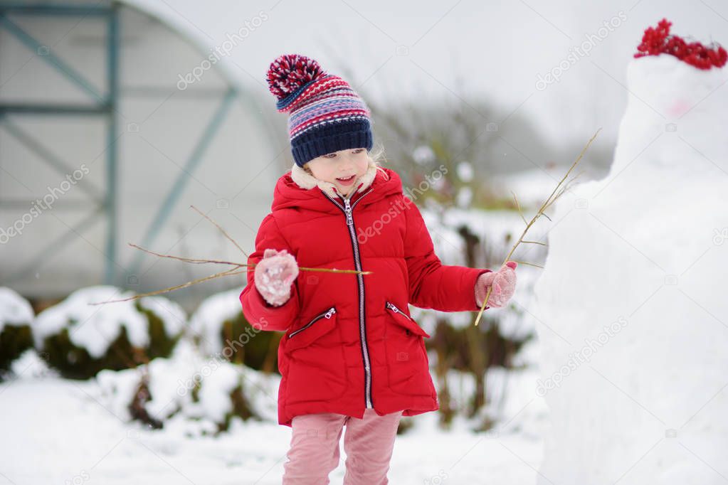 Adorable little girl building a snowman in the backyard. Cute child playing in a snow. Winter activities for kids.