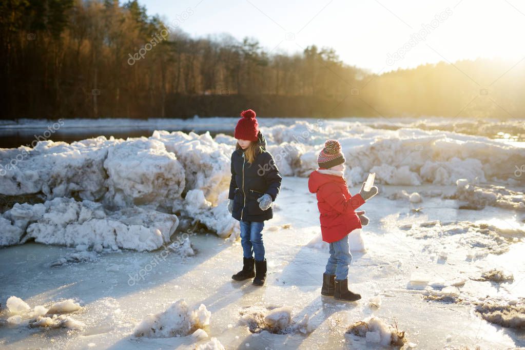 Two happy sisters playing with ice blocks by frozen river during an ice break. Children having fun in winter. Winter activities for kids.