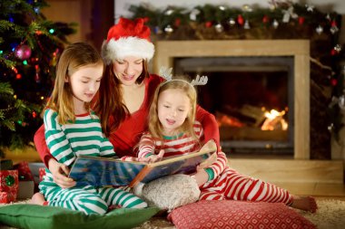 genç anne ve iki küçük kızları kışın rahat oturma odasında bir Noel ağacı tarafından bir kitap okuma
