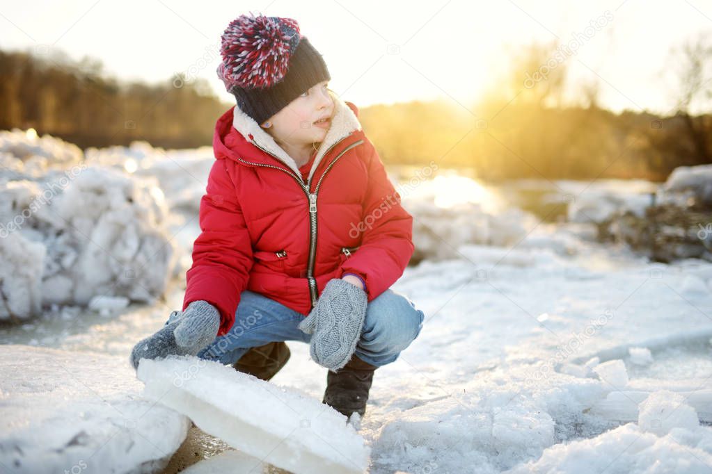 Happy little girl playing with ice blocks by frozen river during an ice break. Child having fun in winter. Winter activities for kids.