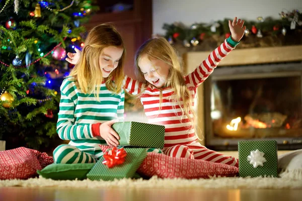 Happy little sisters wearing Christmas pajamas opening gift boxes by a fireplace in a cozy dark living room on Christmas eve. Celebrating Xmas at home.