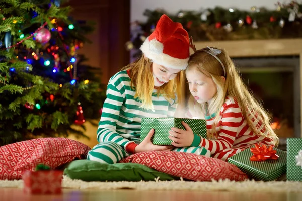 Happy little sisters wearing Christmas pajamas opening gift boxes by a fireplace in a cozy dark living room on Christmas eve. Celebrating Xmas at home.