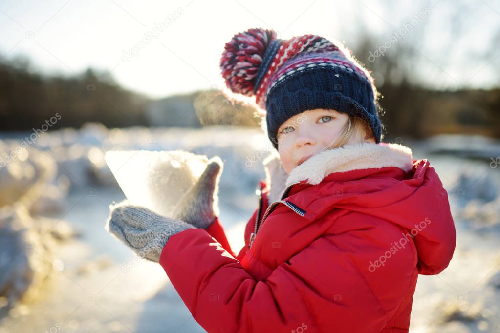 Happy little girl playing with ice blocks by frozen river during an ice break. Child having fun in winter. Winter activities for kids.