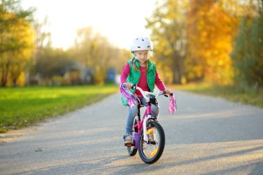 Cute little girl riding a bike in a city park on sunny autumn day. Active family leisure with kids. Child wearing safety hemet while riding a bicycle. clipart
