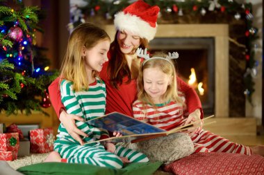 genç anne ve iki küçük kızları kışın rahat oturma odasında bir Noel ağacı tarafından bir kitap okuma