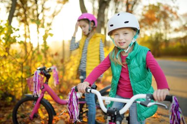Cute little sisters riding bikes in a city park on sunny autumn day. Active family leisure with kids. Children wearing safety hemet while riding a bicycle. clipart