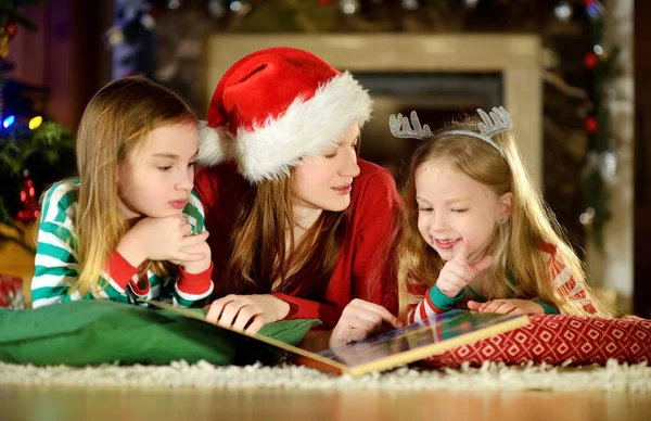 年轻的母亲和两个小女儿读一本书由在舒适的客厅里的一棵圣诞树在冬天 — 图库照片