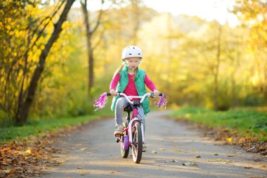 Cute little girl riding a bike in a city park on sunny autumn day. Active family leisure with kids. Child wearing safety hemet while riding a bicycle. clipart