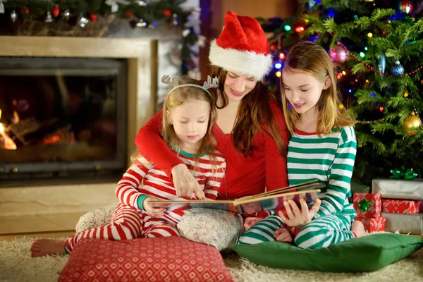 年轻的母亲和她的两个小女儿在舒适的客厅里在圣诞树旁看书 — 图库照片