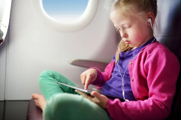 Adorable little girl traveling by an airplane. Child sitting by aircraft window and using a digital tablet during the flight. Traveling abroad with kids.