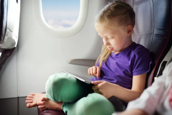 Adorable little girl traveling by an airplane. Child sitting by aircraft window and using a digital tablet during the flight. Traveling abroad with kids.