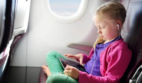 Adorable little girl traveling by an airplane. Child sitting by aircraft window and using a digital tablet during the flight. Traveling abroad with kids.