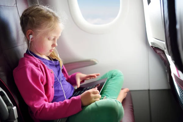 Adorable little girl traveling by an airplane. Child sitting by aircraft window and using a digital tablet during the flight. Traveling abroad with kids.