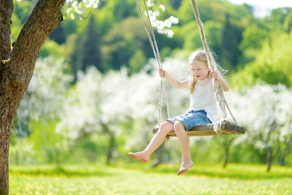 Cute little girl having fun on a swing in blossoming old apple tree garden outdoors on sunny spring day. Spring outdoor activities for kids.