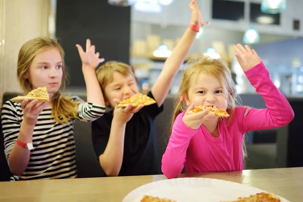 Tři krásné srandovní sourozenci jedí plátky pizzy v domácí restauraci nebo kavárně. — Stock fotografie