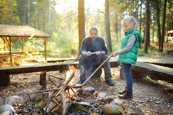Cute young girl learning to start a bonfire. Father teaching her daughter to make a fire. Child having fun at camp fire. Camping with kids in fall forest. — Stock Photo, Image