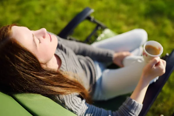 Vrouw ontspannen met gesloten ogen en kopje koffie in lounge stoel op een zonnige dag buitenshuis. — Stockfoto