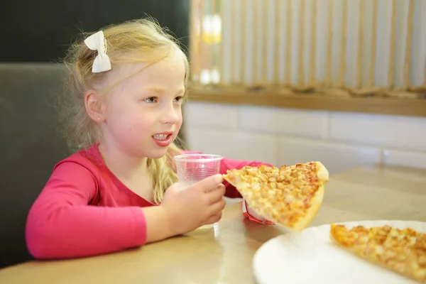Nettes lustiges kleines Mädchen isst ein Stück Pizza im Restaurant oder Café. — Stockfoto